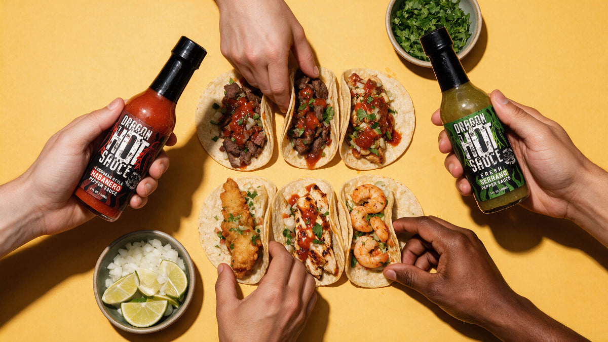 Overhead view of six street tacos on a yellow table as hands reach in, one holding Caribbean Style Habanero and one Fresh Serrano, with bowls of onion, lime and cilantro.