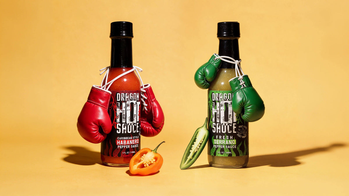 Dragon Caribbean Style Habanero and Fresh Serrano bottles squared off like boxers, wearing tiny red and green boxing gloves, with a halved habanero and serrano at their feet.
