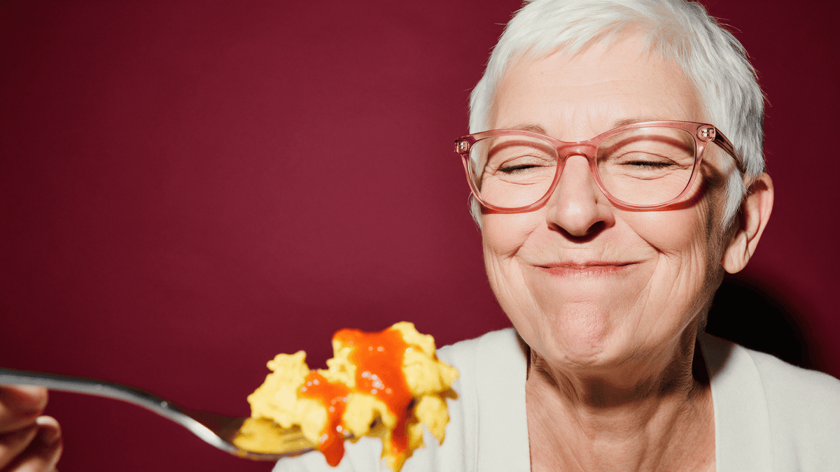 Woman with short white hair and pink glasses savoring a forkful of scrambled eggs with red habanero hot sauce