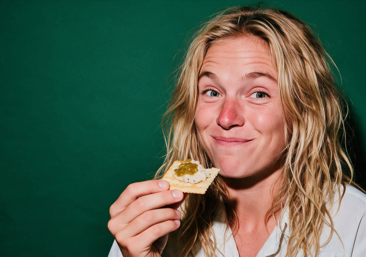Woman with long wavy hair smiling over a cracker topped with fish dip and green Fresh Serrano hot sauce