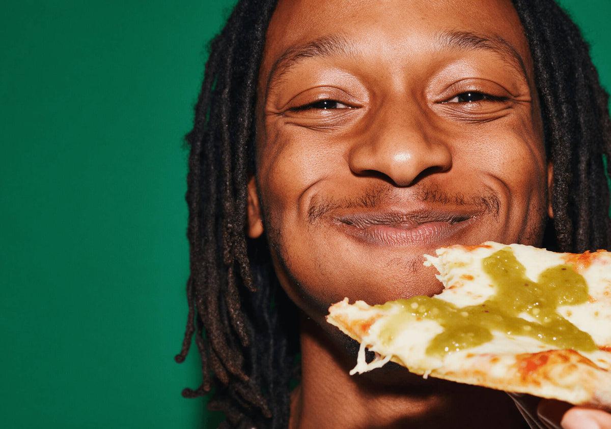 Grinning man holding a slice of pizza topped with green Fresh Serrano hot sauce