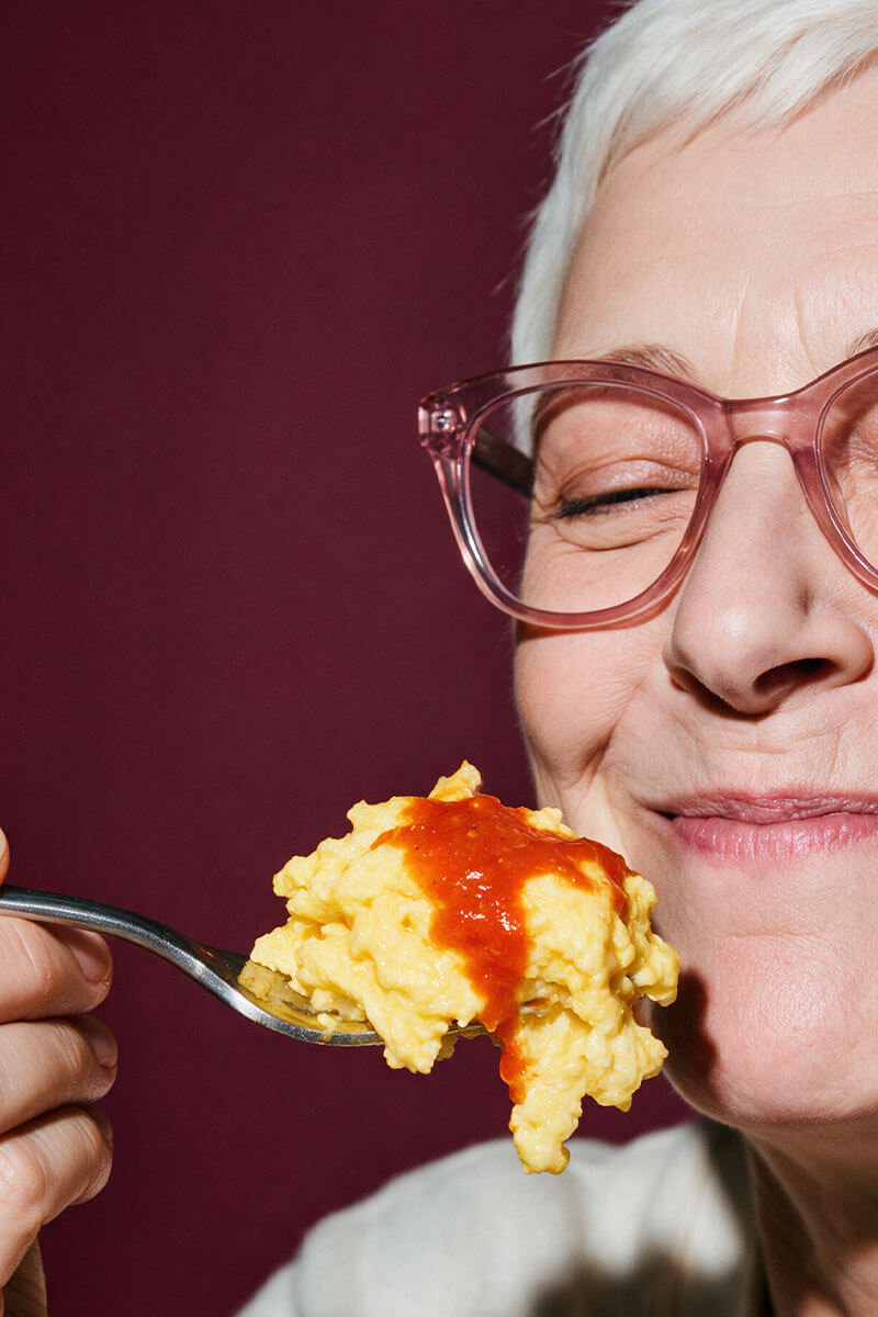 Woman with short white hair and pink glasses savoring a forkful of scrambled eggs with red habanero hot sauce