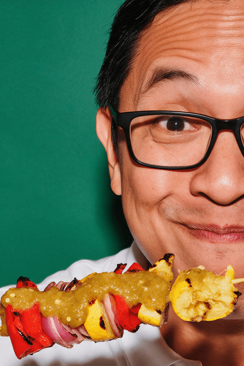 Man in glasses grinning behind a grilled vegetable kabob drizzled with green Fresh Serrano hot sauce
