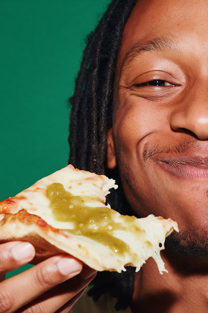 Grinning man holding a slice of pizza topped with green Fresh Serrano hot sauce